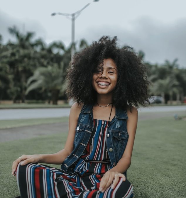 Smiling woman with natural curly hair sitting on grass in San Antonio park wearing denim vest.