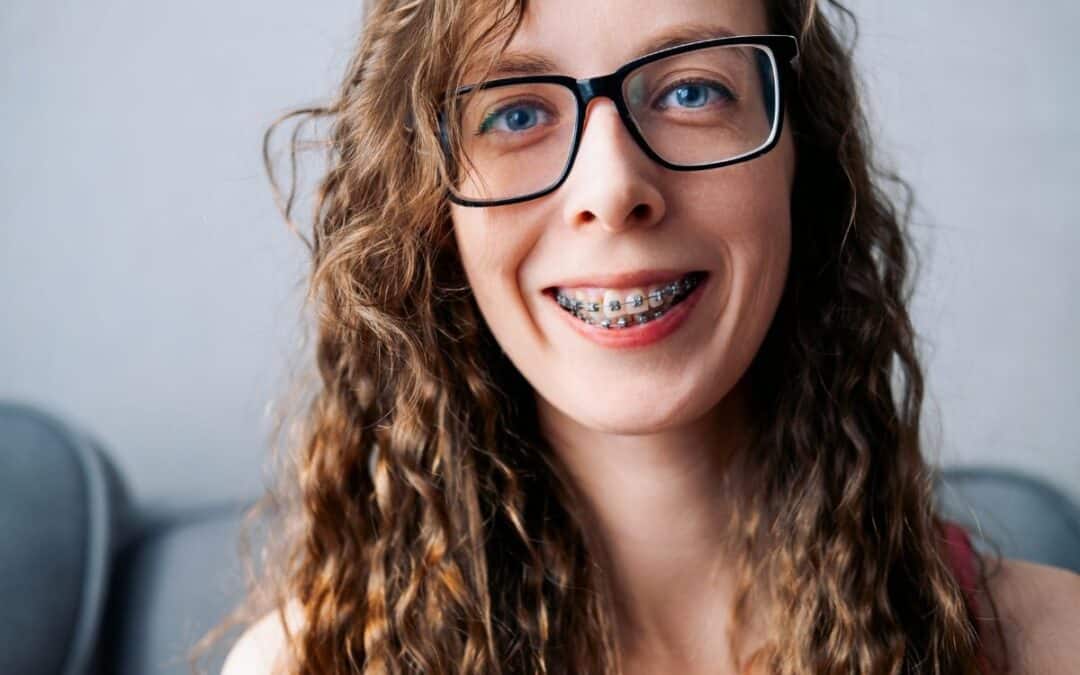 Young woman with curly brown hair and braces smiling at camera wearing glasses.